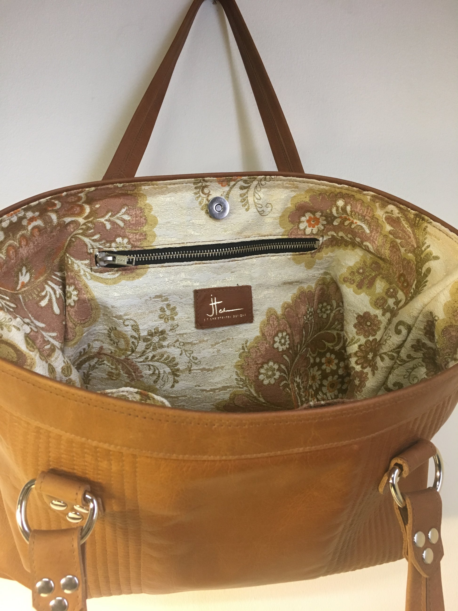 Open Tote with Mercury Style Pleating in Limited Edition Cognac Leather Lined with Re-Purposed 70’s Floral Fabric Lining. Measuring 15” across bottom (19” across top) x 10” x 5” (38 cm At Bottom / 48.25 cm At Top x 25.5 cm Tall x 12.5 cm Wide) and 24” (61cm) Straps. An open divided pocket and zipper pocket with hidden serial number and signature J.T. Christensen Label.